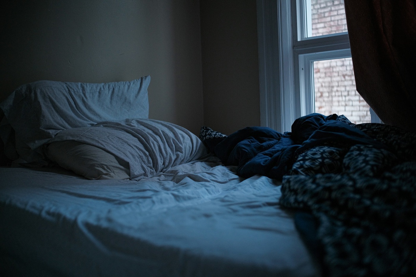 An unmade bed in a dim bedroom, pillows against the wall and a dark patterned duvet pushed aside beneath a window.