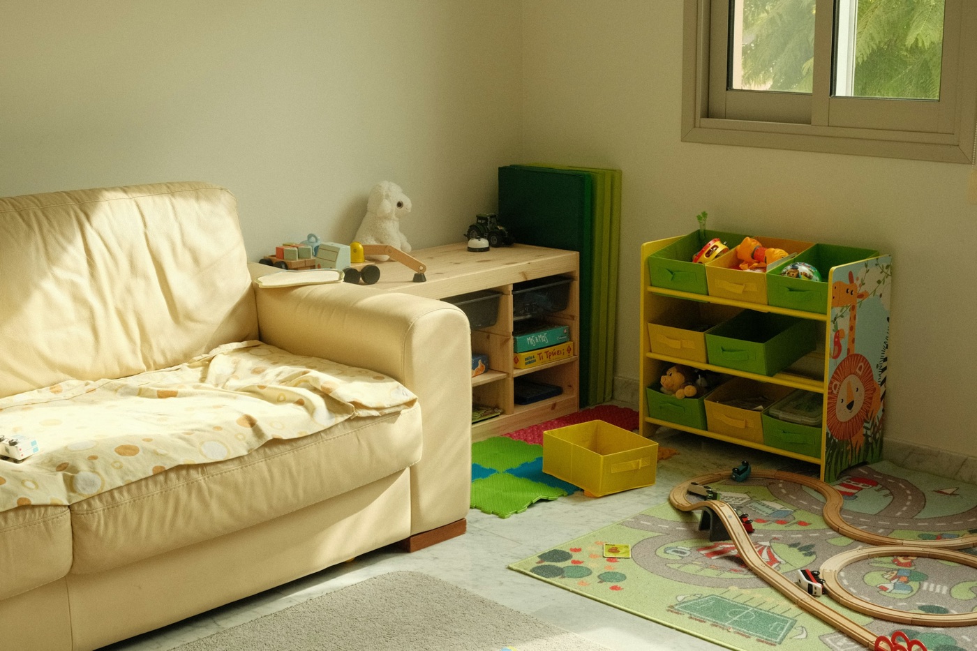 A sunlit living room with a cream sofa, a wooden train track on a play mat, and open toy bins on a low shelf.
