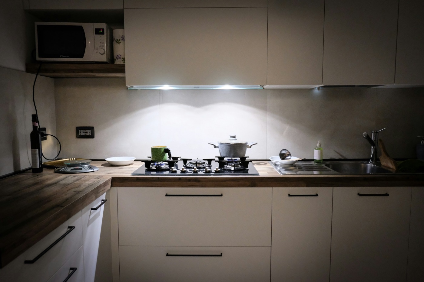 A home kitchen late in the evening, lit only by the light under the cabinets, with a pot on the hob and a nearly clear counter.