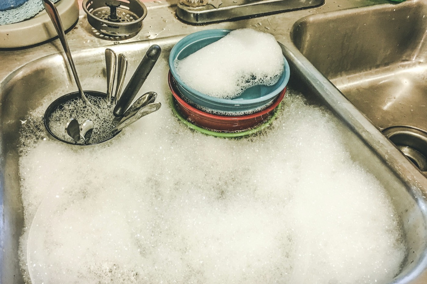 A steel kitchen sink full of soapy water, with a stack of bowls and a cutlery basket soaking in the foam.