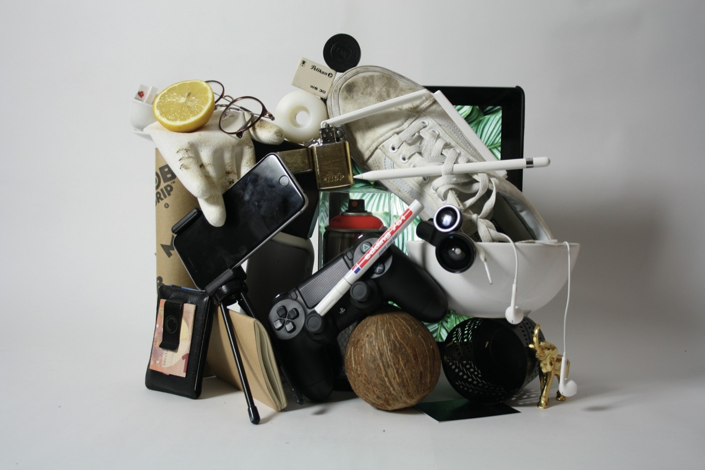 A heap of unrelated things against a white wall: half a lemon, glasses, a phone on a tripod, a sneaker, a game controller, earphones, a wallet and a coconut.