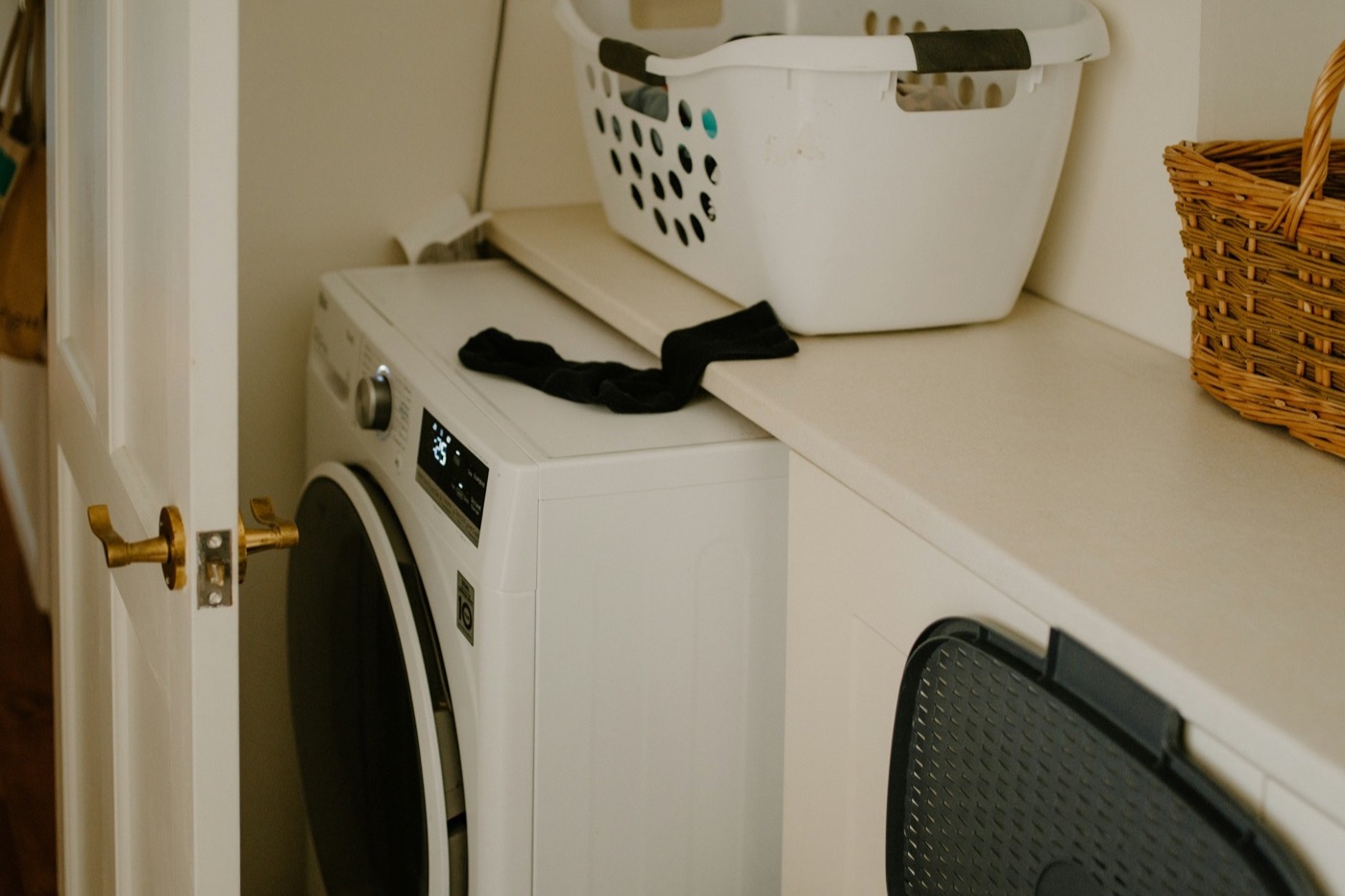 A white front-loading washing machine with a black sock left on top, next to a white plastic laundry basket and a wicker basket on the counter.