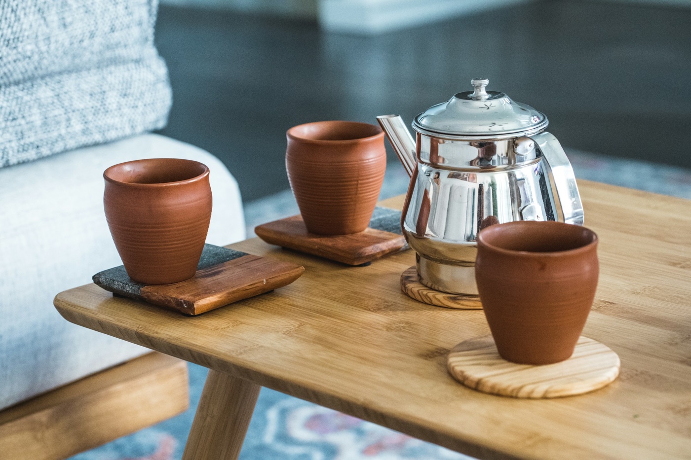 A steel teapot and three terracotta cups on wooden coasters on a low wooden table, next to a grey sofa.