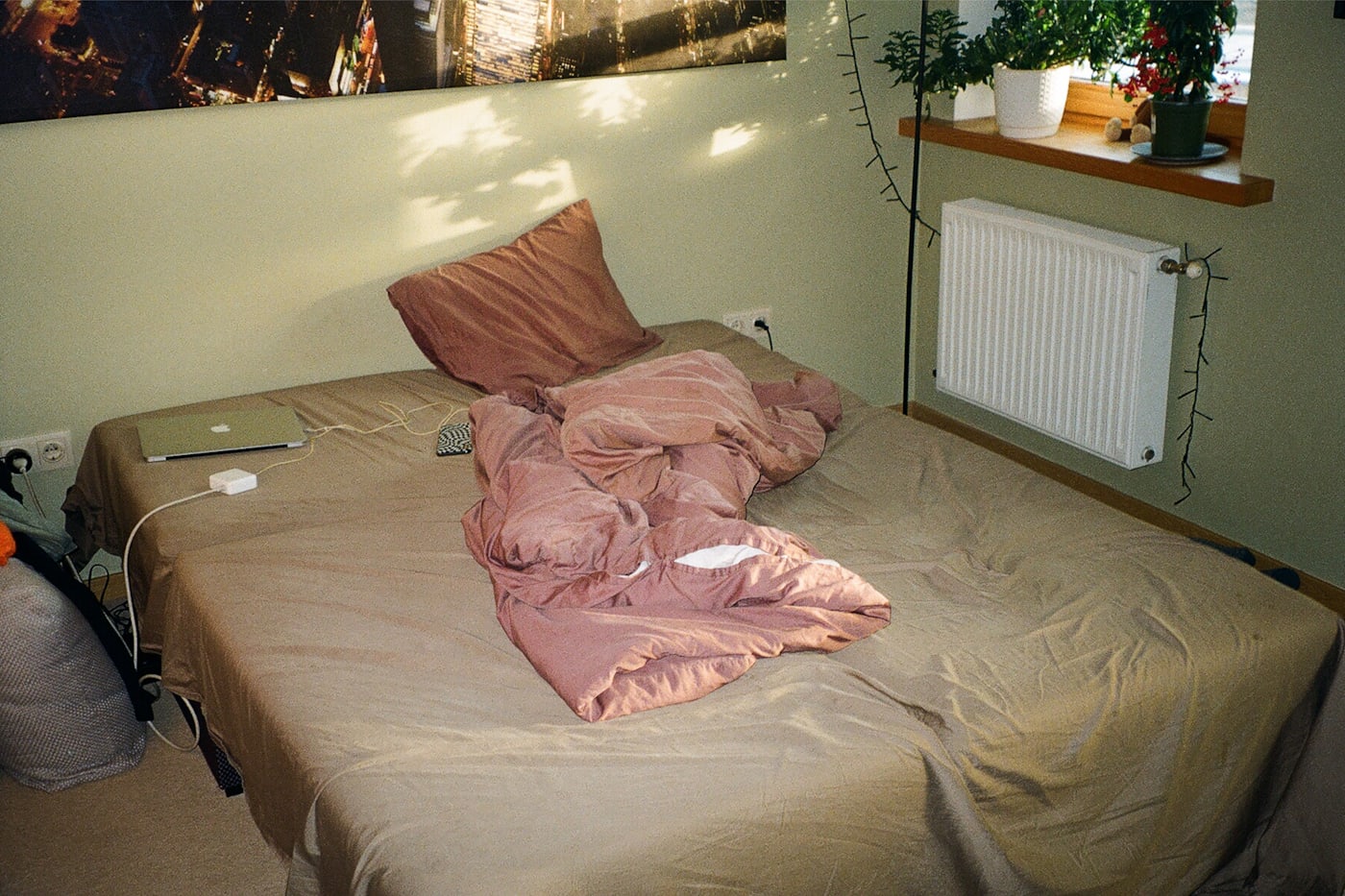 An unmade bed in a small bedroom, duvet pushed to one side, morning light falling across the wall.